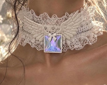 Op de afbeelding: Een witte kanten choker met een centraal vierkant iriserend hangertje met een klein strikje. De choker is versierd met glinsterende witte engelvleugels aan weerszijden en kleine zilveren studs langs de kanten band voor extra textuur.