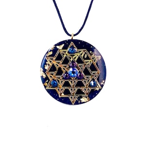 Pode incluir: Um colar com pingente redondo com fundo azul escuro, design geométrico dourado e pedras preciosas azuis e roxas. Detalhes em folha de ouro adornam o pingente. O cordão do colar é azul escuro.