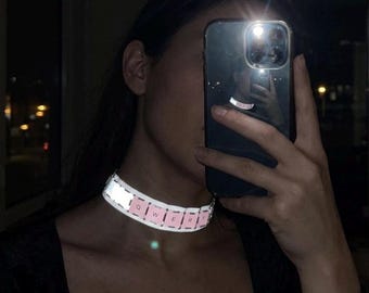 Op de afbeelding: Een reflecterende choker-halsketting met een reeks lichtroze vierkante segmenten, elk bedrukt met een enkele witte letter. De zichtbare letters op de choker spellen QWERT en het materiaal heeft een reflecterende afwerking die oplicht.