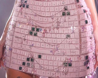 Op de afbeelding: Een lichtroze minirok gemaakt van individuele computertoetsenbordtoetsen. De rok heeft een tailleband van metallic roze rechthoekige schakels en is gedrapeerd met fijne zilveren kettinkjes versierd met kleine bedeltjes, waaronder vlinders, strikjes en rozen.
