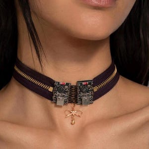 Könnte beinhalten: Ein schwarzes Reißverschluss-Halsband mit zwei kleinen schwarzen Leiterplatten, die silberne USB-Anschlüsse aufweisen und durch eine dunkle Schnürung verbunden sind. Ein zarter, goldfarbener Metallanhänger in Schleifenform hängt in der Mitte.