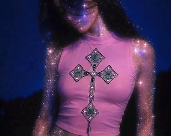 Op de afbeelding: Een mouwloze, lichtroze crop top met een hoge halslijn en een grote, centrale kruisvormige afbeelding. Het kruis is gedetailleerd met donkere omlijningen, groene rechthoekige accenten en heldere, gefacetteerde versieringen in een geometrisch patroon.