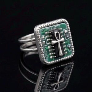 Pode incluir: Anel de prata com um design quadrado de placa de circuito verde e um símbolo ankh preto. O anel tem uma banda dupla. O texto "VOL 707" é visível na placa de circuito.