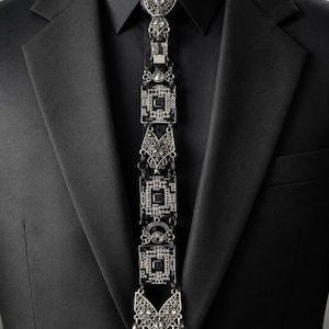 May include: A black suit jacket and shirt worn with a decorative necktie accessory. The tie is constructed from a series of interconnected black and silver metallic circuit board segments, embellished with clear crystals and intricate filigree metalwork.