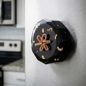 Pode incluir: Um íman de frigorífico preto facetado com detalhes em folha de ouro e um design em forma de flor cor de cobre. O íman está preso à porta de um frigorífico de aço inoxidável numa cozinha.