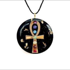 Pode incluir: Um colar com pingente preto com um símbolo ankh dourado adornado com pedras preciosas coloridas. O pingente é circular com detalhes em folha de ouro e pende de um cordão preto.
