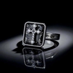 Pode incluir: Anel preto com uma face quadrada com um design de cruz. A cruz é adornada com pequenas pedras brilhantes. O anel tem uma banda de metal escuro e uma superfície refletora.