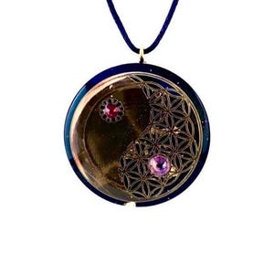 Pode incluir: Um colar com pingente redondo com um cordão azul escuro. O pingente tem um design dourado com um padrão de flor da vida e duas pedras preciosas rosa. O pingente é uma mistura de ouro, azul e rosa.