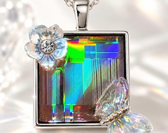 Op de afbeelding: Een zilverkleurige vierkante hanger aan een ketting met een centraal iriserend paneel met verticale lijnen. De hanger is versierd met een doorschijnende, lichtreflecterende bloem met een heldere steen in het midden en een bijpassende vlinder.
