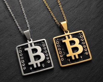 Op de afbeelding: Twee vierkante hangers aan dunne metalen kettingen. De ene hanger is zilverkleurig en de andere goudkleurig. Elk heeft een centraal Bitcoin-symbool tegen een zwarte vierkante achtergrond met kleine cirkelvormige details.
