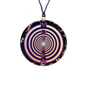 Pode incluir: Um pingente circular com um design em espiral dourado e roxo, adornado com pedras preciosas roxas escuras. O pingente é suspenso por um cordão azul escuro com um fecho dourado. A borda externa é preta com flocos dourados.
