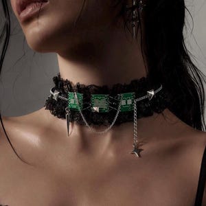 Könnte beinhalten: Ein schwarzes Spitzenhalsband mit drei rechteckigen grünen Leiterplattenkomponenten. Das Schmuckstück ist mit silberfarbenen Metallketten, kleinen pyramidenförmigen Nieten, einem hängenden Dornenanhänger und einem vierzackigen Stern verziert.