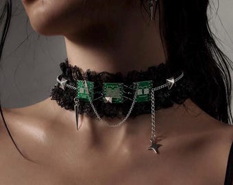 Op de afbeelding: Een zwarte kanten choker met drie rechthoekige groene printplaatcomponenten. Het sieraad is versierd met zilverkleurige metalen kettingen, kleine piramidevormige studs, een bungelende puntige bedel en een vierpuntige ster.