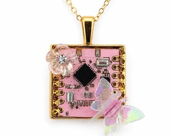 Op de afbeelding: Een goudkleurige ketting met een vierkante hanger gemaakt van een roze printplaat. De hanger is versierd met een doorzichtige, iriserende bloem met een centrale strass-steen en een doorschijnende, iriserende vlinderfiguur aan de zijkant.