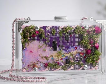 Op de afbeelding: Een rechthoekige doorzichtige clutch van acryl met een paarse printplaat aan de binnenkant, versierd met gedroogd groen mos, kleine roze bloemen en verspreide blauwe en zilveren strass-steentjes. De tas heeft zilveren metalen hoeken en een roze ketting.