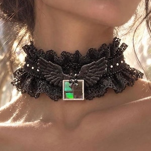 May include: A black lace choker necklace featuring a ruffled edge and a central silver-toned charm. The charm consists of a pair of dark wings positioned above a square pendant with a small bow and a shimmering, iridescent green and black inlay.