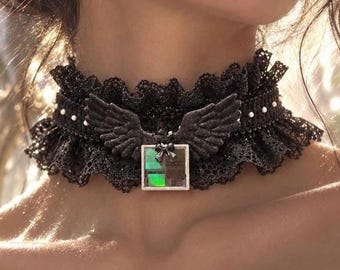 Op de afbeelding: Een zwarte kanten choker met een geplooide rand en een centrale zilverkleurige bedel. De bedel bestaat uit een paar donkere vleugels boven een vierkante hanger met een kleine strik en een glinsterende, iriserende groen-zwarte inleg.