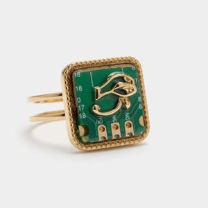 Pode incluir: Anel dourado com um design quadrado de placa de circuito verde e um símbolo dourado do Olho de Hórus. O anel tem uma banda dupla e uma borda texturizada. Uma joia única.