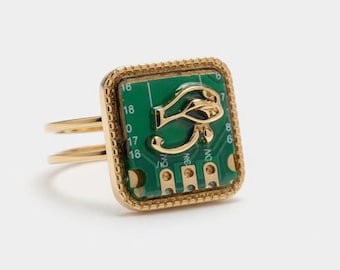 Op de afbeelding: Gouden ring met een vierkant groen printplaatontwerp en een gouden Eye of Horus-symbool. De ring heeft een dubbele band en een getextureerde rand. Een uniek sieraad.
