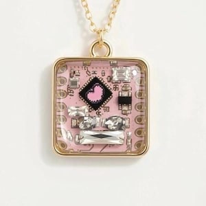 Op de afbeelding: Gouden ketting met een vierkante hanger. De hanger heeft een roze printplaatontwerp, een gouden frame, heldere kristallen en een zwart vierkant met een roze hart.