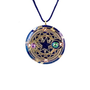 Pode incluir: Pingente redondo azul escuro com um design dourado de estrela e detalhes dourados. O pingente tem uma argola dourada e um cordão azul escuro. É adornado com pedras preciosas rosa e verdes.