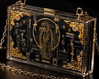 Op de afbeelding: Een rechthoekige box-clutch van helder acryl met een zwart printplaat-interieur en een goudkleurige afbeelding van de Maagd Maria. De buitenkant is versierd met sierlijke goudkleurige metalen hoekstukken, kleine kruisbedels en een gouden ketting.