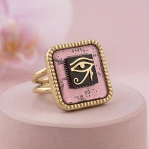 Pode incluir: Anel dourado com um design quadrado rosa e preto do Olho de Hórus. O anel tem uma banda dupla e uma borda decorativa. O Olho de Hórus é um símbolo egípcio de proteção e poder real.