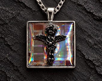 Op de afbeelding: Een zilverkleurige vierkante hanger aan een ketting met een centrale donker metalen cherubijnfiguur. De achtergrond van de hanger is een reflecterend, iriserend printplaatpatroon met regenboogkleurige lichtbrekingen, hangend aan een zilverkleurige ketting.