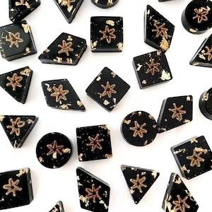 Pode incluir: Uma coleção de objetos decorativos pretos de formato geométrico com detalhes em folha de ouro. Cada peça apresenta um design de estrela dourada. As formas incluem quadrados, triângulos, losangos e círculos, sobre um fundo branco.