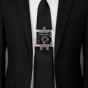 May include: A black necktie featuring a central electronic circuit board component integrated into the fabric. The rectangular black circuit board is attached to the upper and lower sections of the tie with silver-toned metal connectors.