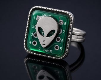 Op de afbeelding: Een vierkante ring met een groene printplaat als achtergrond en een zilverkleurig alienhoofd in het midden. De printplaat toont de cijfers één tot en met acht en de tekst O. ZAMM. De ring heeft een gestructureerde zilverkleurige metalen band.