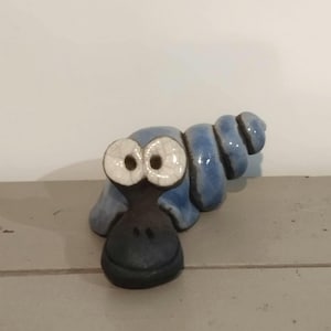 Peut inclure: Une figurine d'escargot en céramique avec une coquille en spirale bleu brillant et une tête gris foncé. L'escargot possède deux grands yeux blancs proéminents avec une finition craquelée, lui donnant une expression écarquillée.