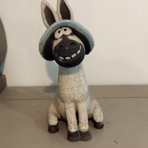Peut inclure: Figurine d'âne en céramique souriant, coiffé d'un chapeau bleu. L'âne est gris clair avec des sabots et des oreilles brun foncé. La figurine présente une finition craquelée, lui donnant un aspect vintage.