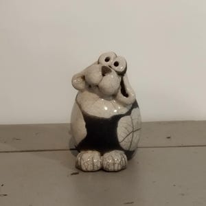 Peut inclure: Une figurine en céramique représentant un chien au corps crème orné d'un motif de glaçure craquelée brun foncé. Le chien possède de grands yeux ronds, un museau proéminent, des oreilles tombantes et un corps arrondi et trapu.