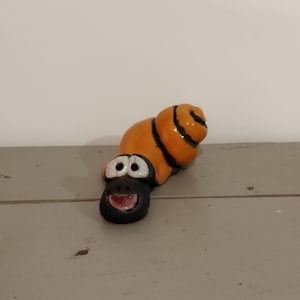 Peut inclure: Une petite figurine d'escargot en céramique avec une coquille orange brillante ornée de spirales brun foncé. L'escargot possède une tête noire avec deux grands yeux blancs, un petit nez et une bouche ouverte révélant un intérieur rose, le tout verni.