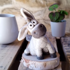 Könnte beinhalten: Eine Esel-Figur aus Keramik mit einer rissigen, cremeweißen Glasur sowie dunkelbrauner Schnauze, Ohren und Hufen. Der Esel hat große, weit auseinanderstehende Augen und sitzt auf einem runden Sockel aus einer hellen Naturholzscheibe.