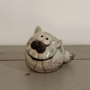 Peut inclure: Une figurine de chat en céramique ronde avec une finition émaillée craquelée blanche. Le chat a deux petits yeux circulaires, un nez arrondi brun foncé avec trois points et des oreilles pointues. Une petite queue dépasse du corps sphérique.