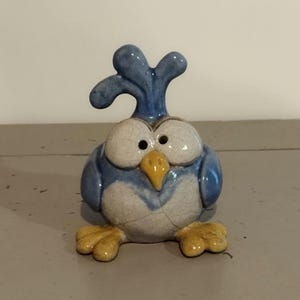 Peut inclure: Une figurine d'oiseau en céramique avec un corps bleu à finition craquelée, de grands yeux blancs à pupilles noires, un bec jaune et des pattes jaunes. L'oiseau possède une crête bleue sur la tête et un corps arrondi et trapu avec de petites ailes.