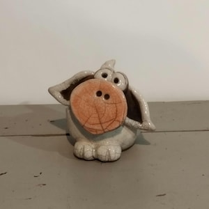 Peut inclure: Figurine de cochon en céramique avec un grand museau orange texturé et de grandes oreilles. Le cochon est principalement blanc avec un effet de glaçure craquelée sur le museau. La figurine a un design fantaisiste et caricatural.