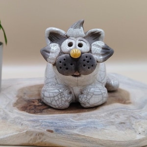 Op de afbeelding: Een keramieken kattenbeeldje met een grillig ontwerp. De kat is voornamelijk wit met grijze accenten op de oren, neus en poten. Het heeft een gele neus en grote, ronde ogen. Het beeldje staat op een houten plank.