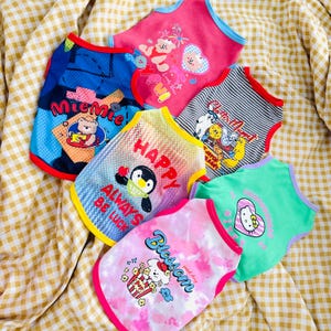 May include: A collection of colorful pet tank tops with various graphic designs. The tops feature images of bears, penguins, and cartoon characters. The tank tops are in shades of pink, blue, green, and white, with contrasting trim.