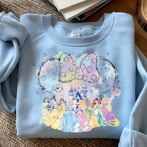 Può includere: Felpa azzurra con una grafica colorata di un fiocco a pois, orecchie di topo e un castello. Sotto, illustrazioni di principesse in abiti. La felpa è realizzata in materiale morbido.