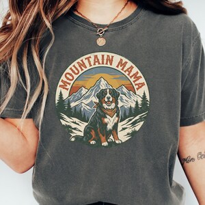 May include: Dark grey t-shirt with a circular graphic. The graphic shows a dog wearing a bandana in front of a mountain range and trees, with the text "MOUNTAIN MAMA." The shirt is made of a soft material.