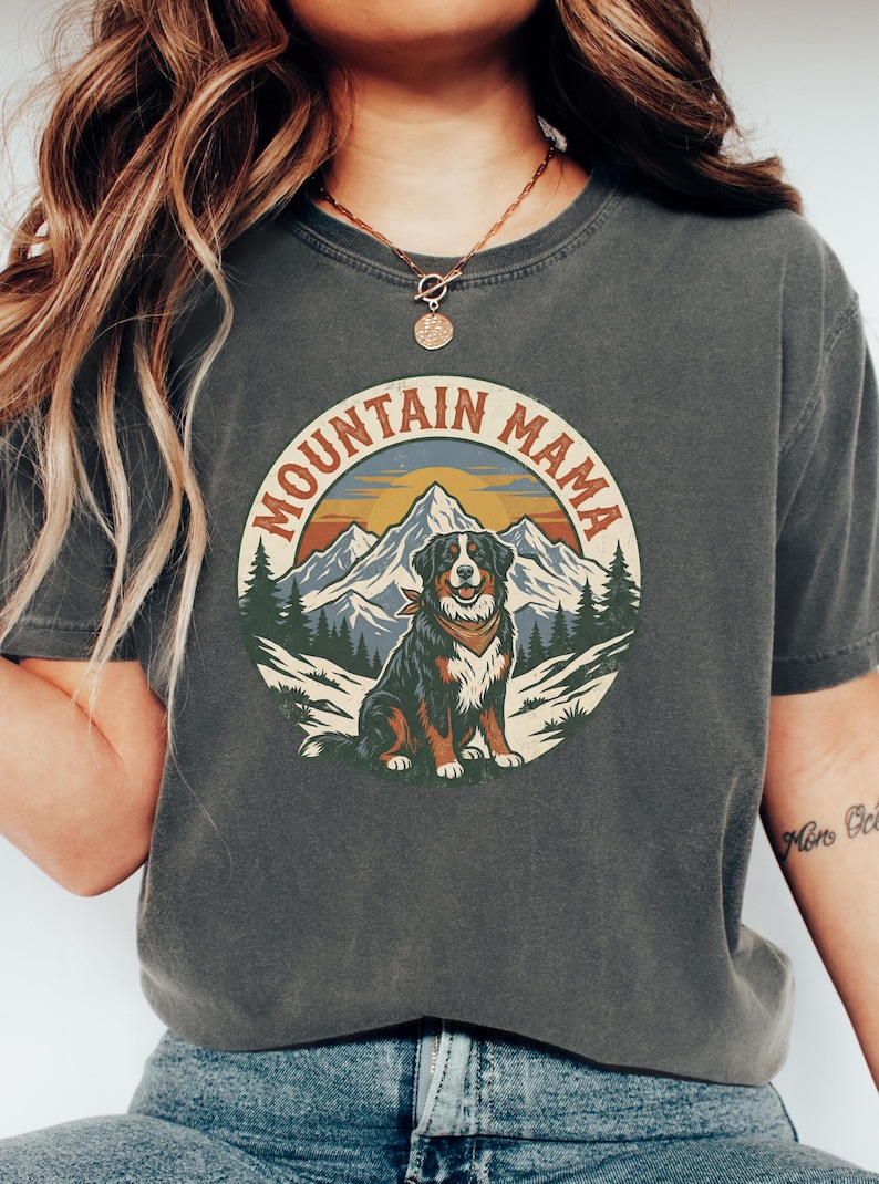 May include: Dark grey t-shirt with a circular graphic. The graphic shows a dog wearing a bandana in front of a mountain range and trees, with the text "MOUNTAIN MAMA." The shirt is made of a soft material.