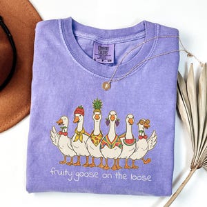 May include: A light purple Comfort Colors t-shirt featuring a graphic of six cartoon geese, each adorned with fruit-themed accessories. The text "fruity goose on the loose" is printed below the geese. A gold necklace and a brown hat are also visible.