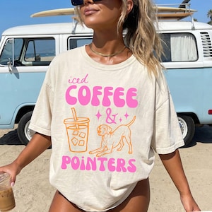 May include: Cream-colored t-shirt with the words "iced COFFEE & POINTERS" in pink and orange. The design includes a graphic of an iced coffee and a dog. The shirt is a casual, oversized fit.