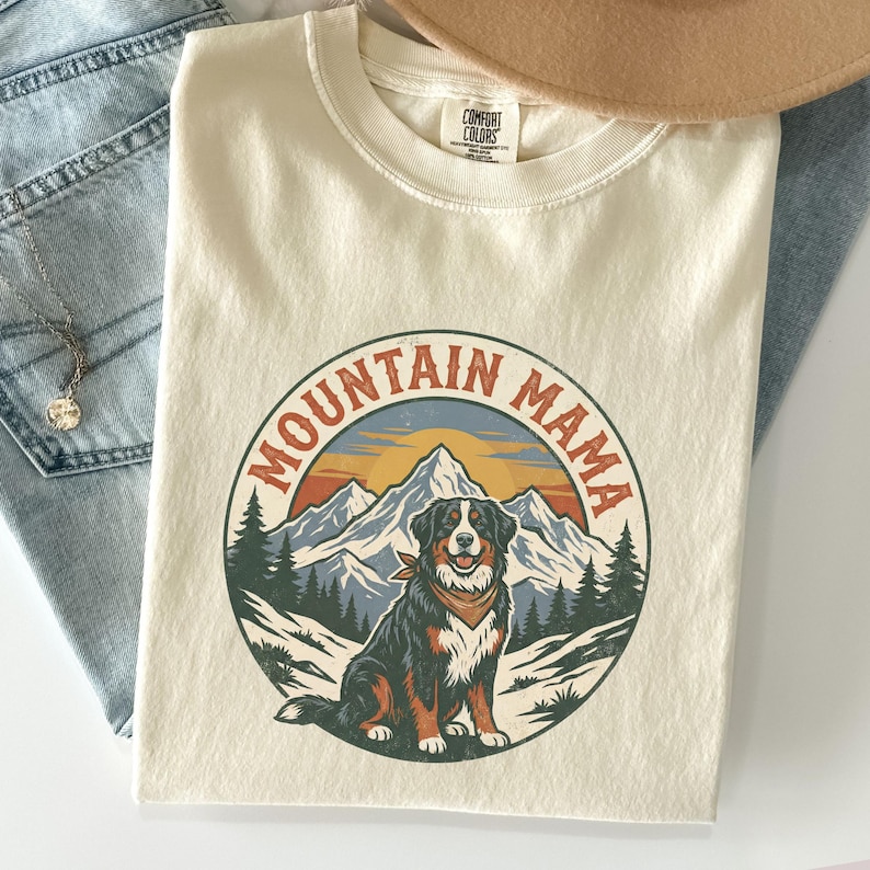May include: Cream-coloured t-shirt with a circular graphic featuring a mountain scene and a dog wearing a bandana. The text "MOUNTAIN MAMA" is arched around the graphic. A tan hat and blue jeans are in the background.