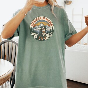 May include: A sage green t-shirt with a circular graphic that reads "MOUNTAIN MAMA". The graphic depicts a dog, mountains, and trees. The shirt has short sleeves and a loose fit.