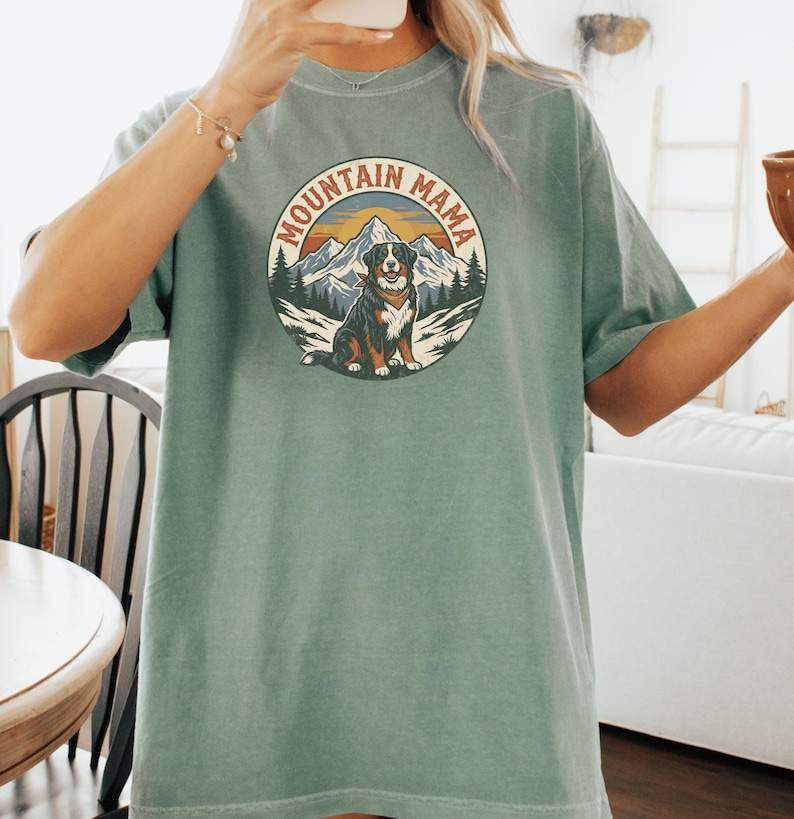 May include: A sage green t-shirt with a circular graphic that reads "MOUNTAIN MAMA". The graphic depicts a dog, mountains, and trees. The shirt has short sleeves and a loose fit.