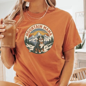 May include: Orange short-sleeved t-shirt with a circular graphic featuring a dog, mountains, and the text "MOUNTAIN MAMA". The dog is brown, black, and white. The shirt is made of a soft material.
